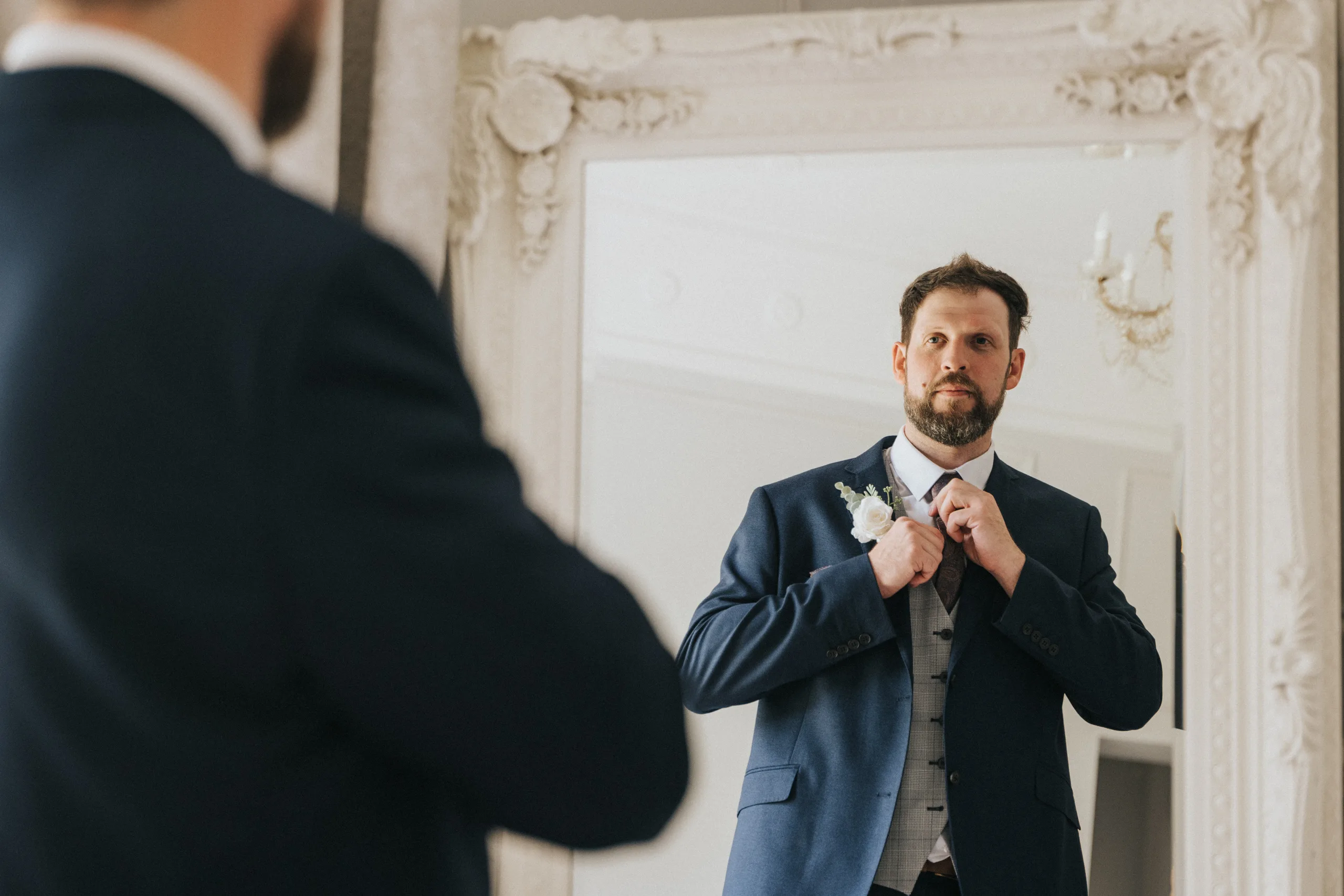 shane-carina-wedding-photography-scarborough-hackness-grange-photographer-copyright-aimee-lince-76-of-649-©-aimee-lince-photography-16-07-2025 A man in a navy suit adjusts his tie while looking at himself in an ornate, white-framed mirror. Preparing for his special day at Hackness Grange, the groom stands poised as a Scarborough wedding photographer captures the moment in a bright, elegant room. © Aimee Lince Photography - Wedding photographer in Lincolnshire, Yorkshire & Nottinghamshire