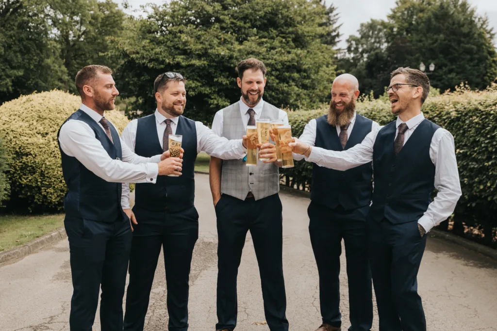 Five men dressed in formal attire, wearing vests and ties, stand outdoors at Hackness Grange near Scarborough. Smiling, they clink large beer glasses together in a celebratory toast, surrounded by lush greenery—perfect for a wedding photographer to capture. © Aimee Lince Photography - Wedding photographer in Lincolnshire, Yorkshire & Nottinghamshire