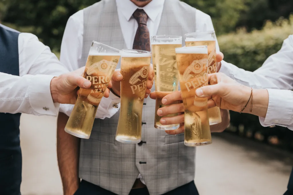Five men in dress shirts and vests clink tall glasses of Peroni beer outdoors at Hackness Grange. Only their torsos and hands are visible, the blurred greenery hinting at a celebratory event—a moment a wedding photographer in Scarborough would love to capture. © Aimee Lince Photography - Wedding photographer in Lincolnshire, Yorkshire & Nottinghamshire