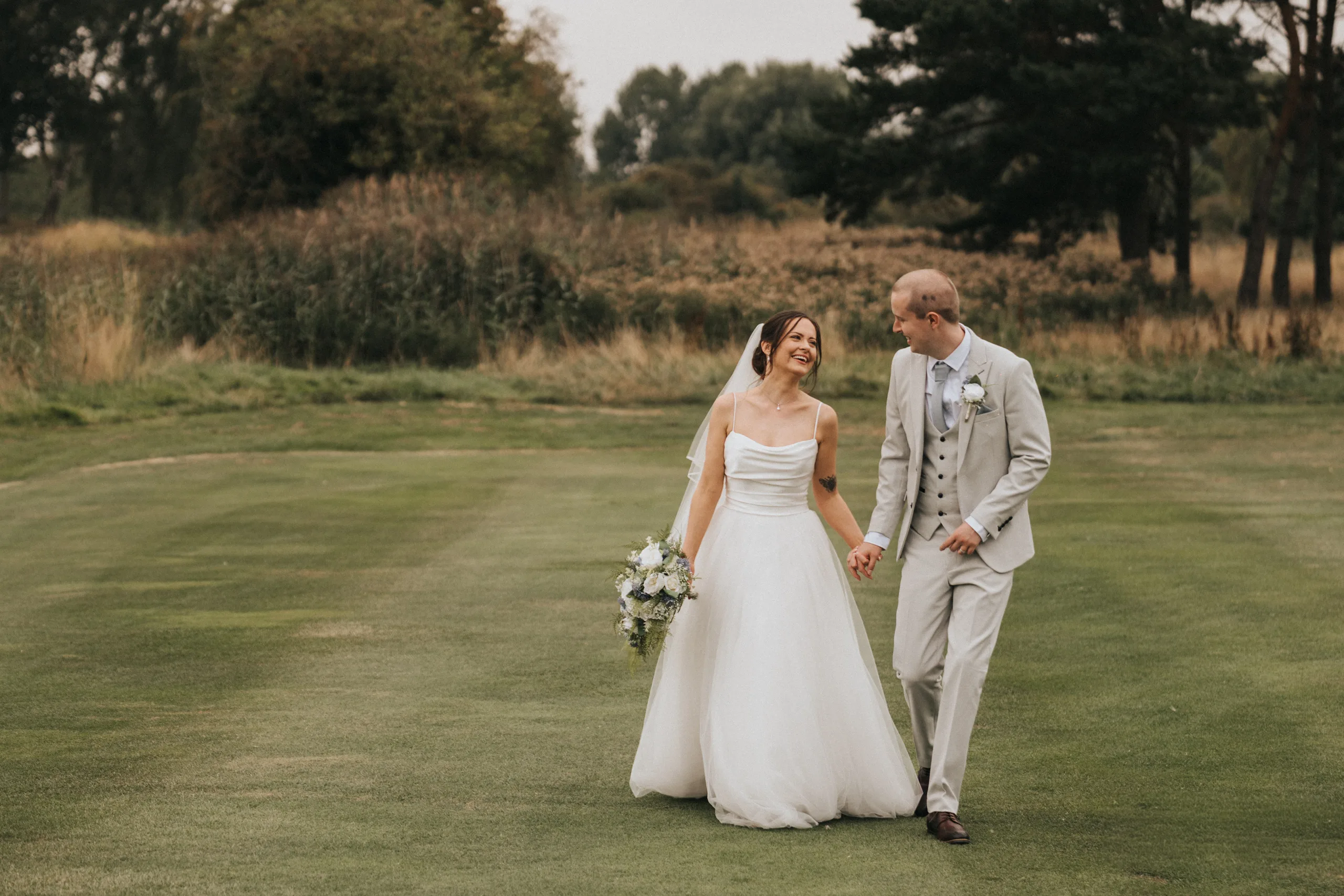 A bride in a white gown holds a bouquet and smiles at her groom as they walk hand in hand across a grassy field—capturing the true wedding photography worth under an overcast sky with trees and bushes in the background. © Aimee Lince Photography - Wedding photographer in Lincolnshire, Yorkshire & Nottinghamshire