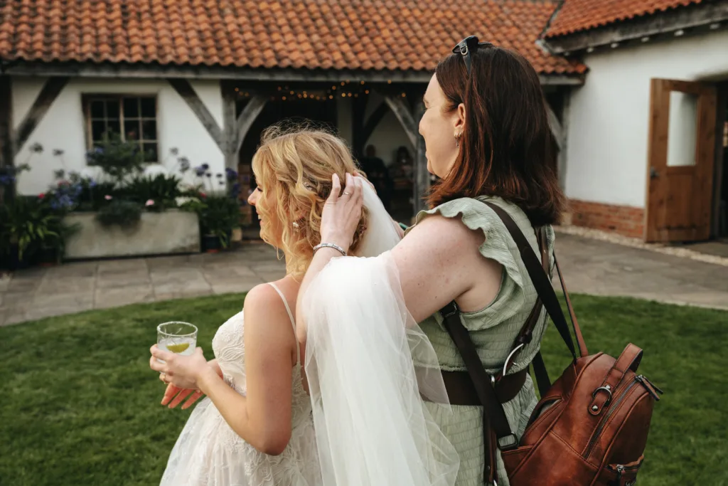 A woman in a green dress adjusts the veil of a bride in a white gown holding a drink. They stand on a lawn in front of a rustic building with white walls, wooden beams, and a tiled roof. The bride faces away; the atmosphere is relaxed and candid. © Aimee Lince Photography - Wedding photographer in Lincolnshire, Yorkshire & Nottinghamshire