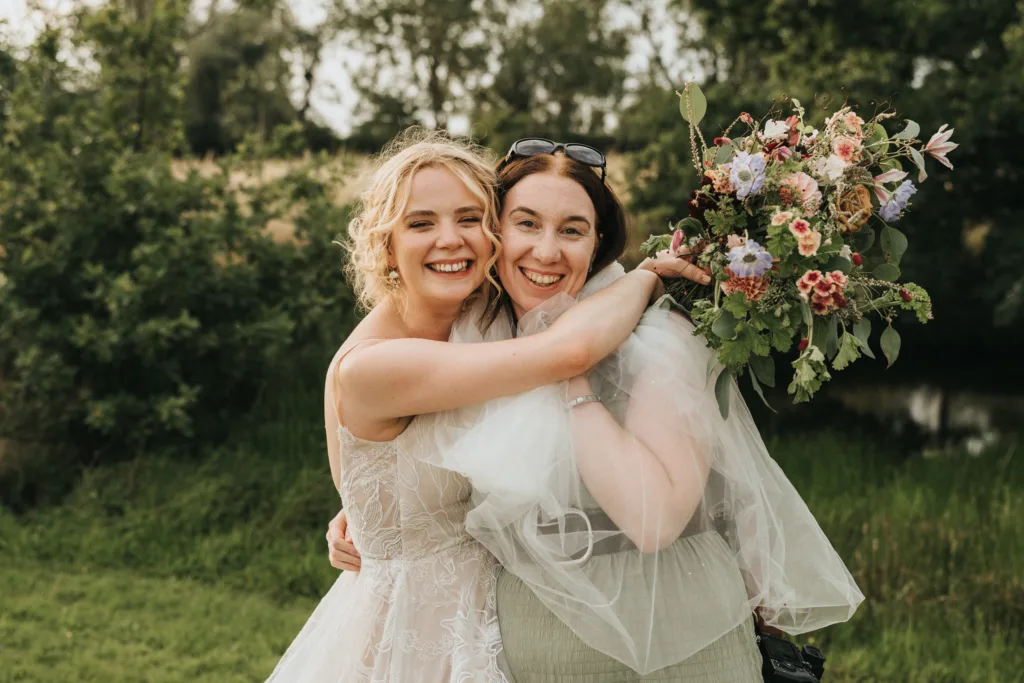 abbey-farm-weddings-behind-the-scenes-1-of-1-©-aimee-lince-photography-09-08-2025-3 Two women smiling and embracing outdoors; one wears a white lace wedding dress and veil, holding a bouquet of colorful flowers, while the other wears a light sage green dress and sunglasses on her head. Lush greenery and trees fill the background. © Aimee Lince Photography - Wedding photographer in Lincolnshire, Yorkshire & Nottinghamshire