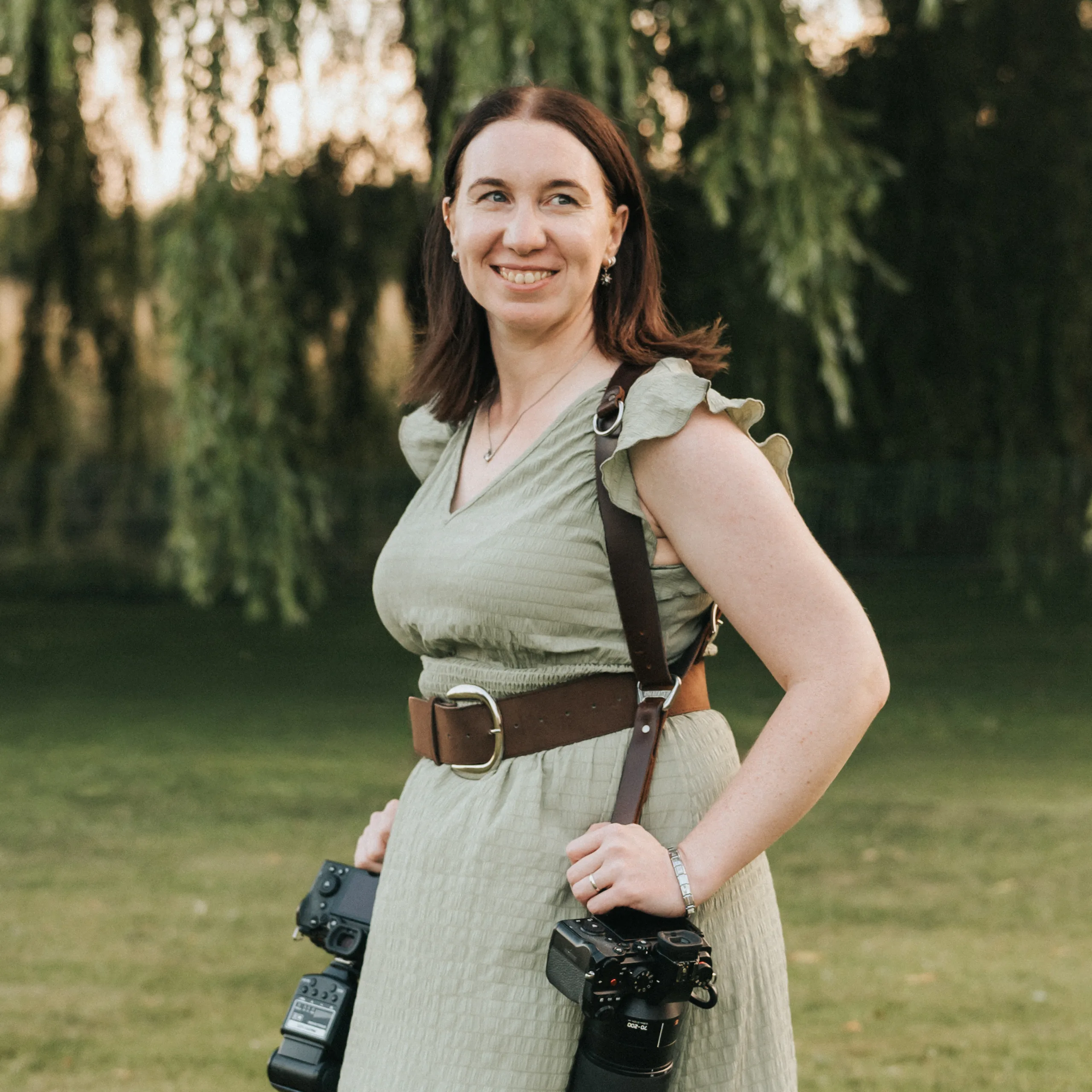A woman with shoulder-length brown hair, wearing a light green dress and brown belt, stands on grass with trees behind her. Smiling, she carries two cameras across her body—ready for wedding photography in the warm late afternoon light. © Aimee Lince Photography - Wedding photographer in Lincolnshire, Yorkshire & Nottinghamshire