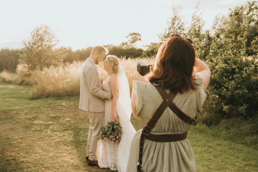 A photographer captures a bride and groom in a sunlit field, embracing lovingly in signature lifestyle fashion. The couple, dressed in wedding attire, is framed amid tall grass and trees bathed in warm light. © Aimee Lince Photography - Wedding photographer in Lincolnshire, Yorkshire & Nottinghamshire