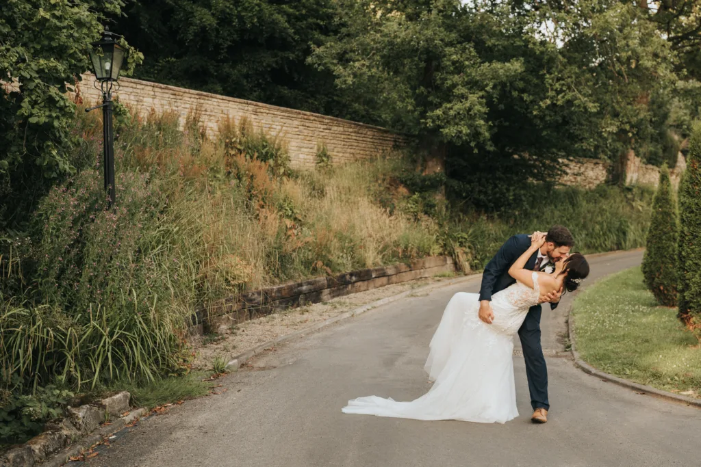 A groom in a navy suit dips and kisses a bride in a white lace wedding dress on a quiet Lincolnshire road, surrounded by lush greenery and a stone wall. This romantic, sunlit moment is wedding photography at its finest, framed by tall grass and trees. © Aimee Lince Photography - Wedding photographer in Lincolnshire, Yorkshire & Nottinghamshire