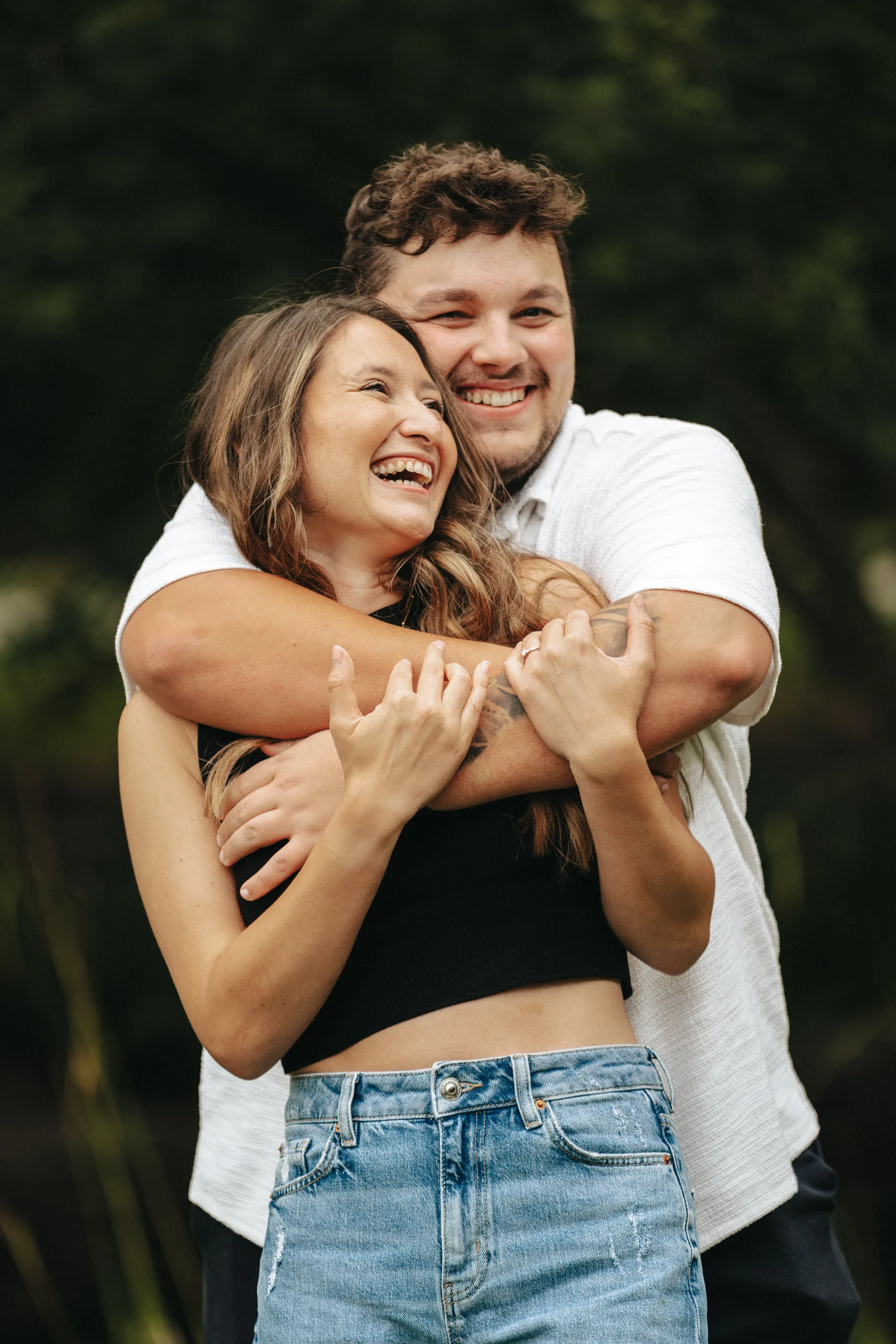 A smiling couple stands outdoors, their affectionate embrace radiating joy—perfect inspiration for wedding photography. The blurred greenery background highlights their happiness, with the man in a white shirt hugging his laughing partner in a black crop top and blue jeans. © Aimee Lince Photography - Wedding photographer in Lincolnshire, Yorkshire & Nottinghamshire