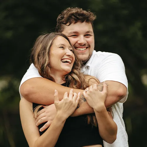 leah-jordan-engagement-pre-wedding-photoshoot-oaklands-wedding-venue-driffield-1-of-2-©-aimee-lince-photography-18-08-2025 A young couple stands outdoors, smiling and laughing. The man, in a white shirt, hugs the woman from behind as she beams with joy—captured perfectly for wedding photography amid green trees in the background. © Aimee Lince Photography - Lincolnshire, Yorkshire & Nottinghamshire