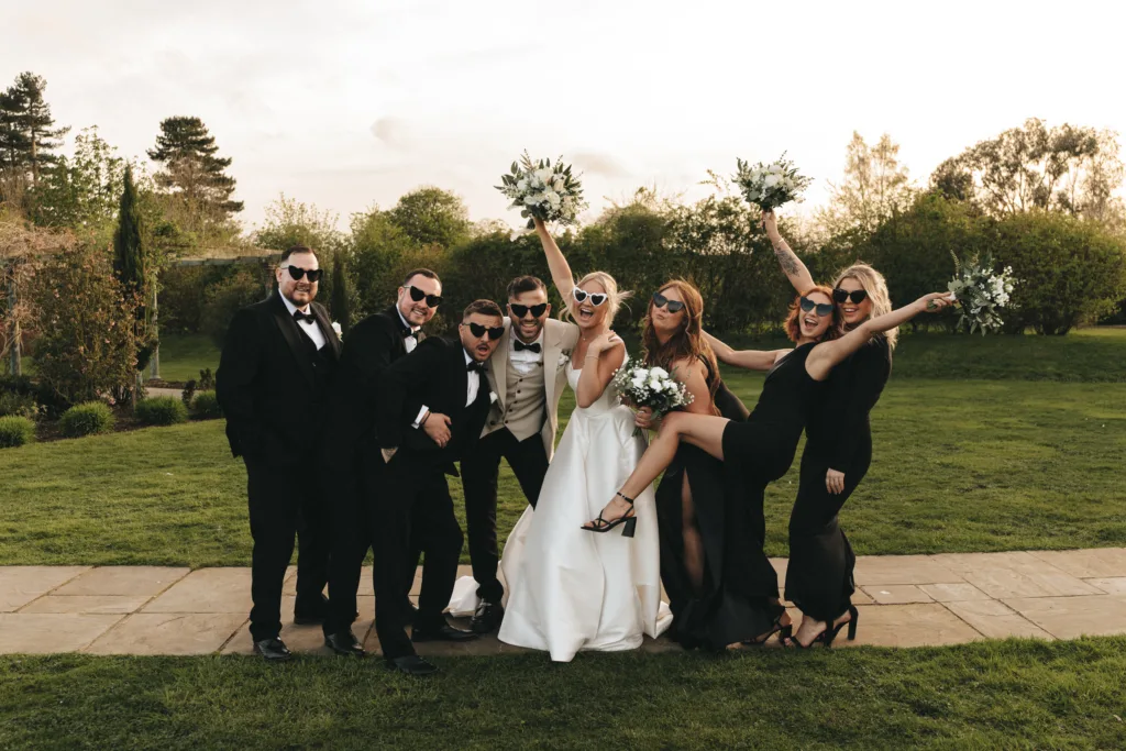 A joyful wedding party poses outdoors on grass at sunset, captured by a talented wedding photographer. The bride in white lifts her leg, surrounded by bridesmaids and groomsmen in black attire—all wearing sunglasses and smiling enthusiastically. © Aimee Lince Photography - Wedding photographer in Lincolnshire, Yorkshire & Nottinghamshire