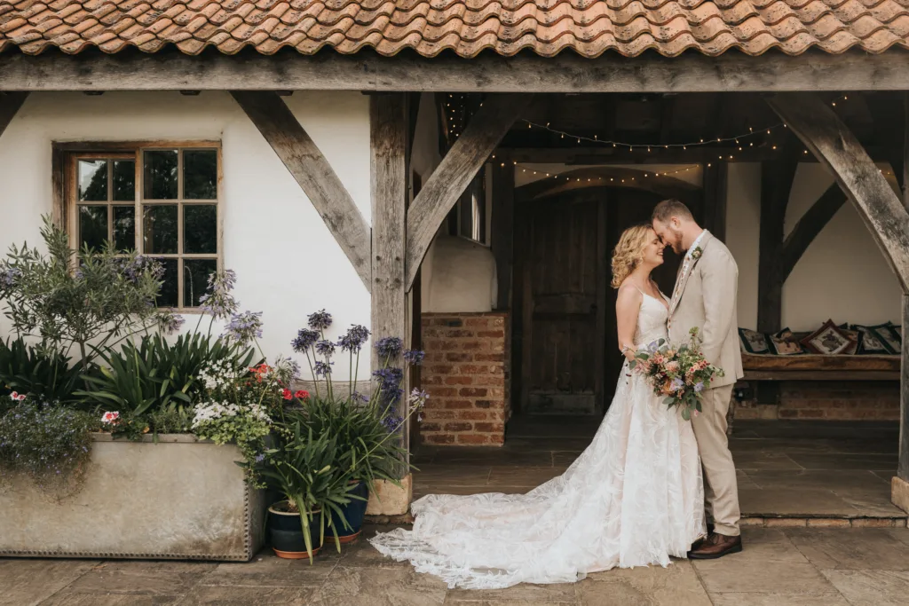 A bride and groom stand close together, foreheads touching, outside a rustic building with wooden beams. String lights and lush flowers frame the romantic scene—an enchanting moment captured by wedding photography. © Aimee Lince Photography - Wedding photographer in Lincolnshire, Yorkshire & Nottinghamshire