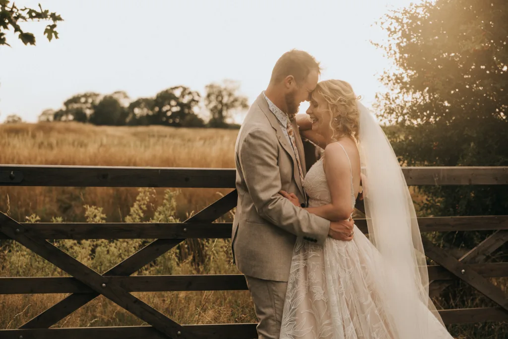 A bride and groom embrace beside a wooden gate in a sunlit field, captured beautifully by a wedding photographer. The couple stands close, smiling tenderly amid golden grass and trees, the bride's lace gown and veil glowing in the light. © Aimee Lince Photography - Wedding photographer in Lincolnshire, Yorkshire & Nottinghamshire