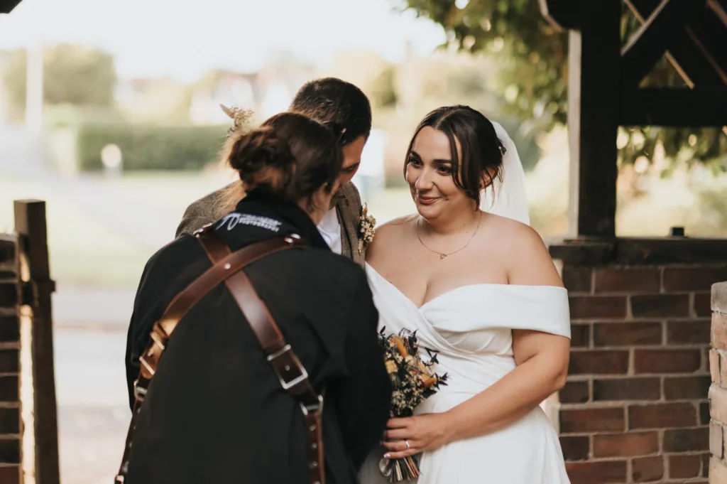 A bride in an off-shoulder white dress and veil smiles, holding a bouquet. A groom stands close beside her, partially obscured. Another person, seen from behind, faces them. They are outdoors near a brick wall with greenery in the background. © Aimee Lince Photography - Wedding photographer in Lincolnshire, Yorkshire & Nottinghamshire