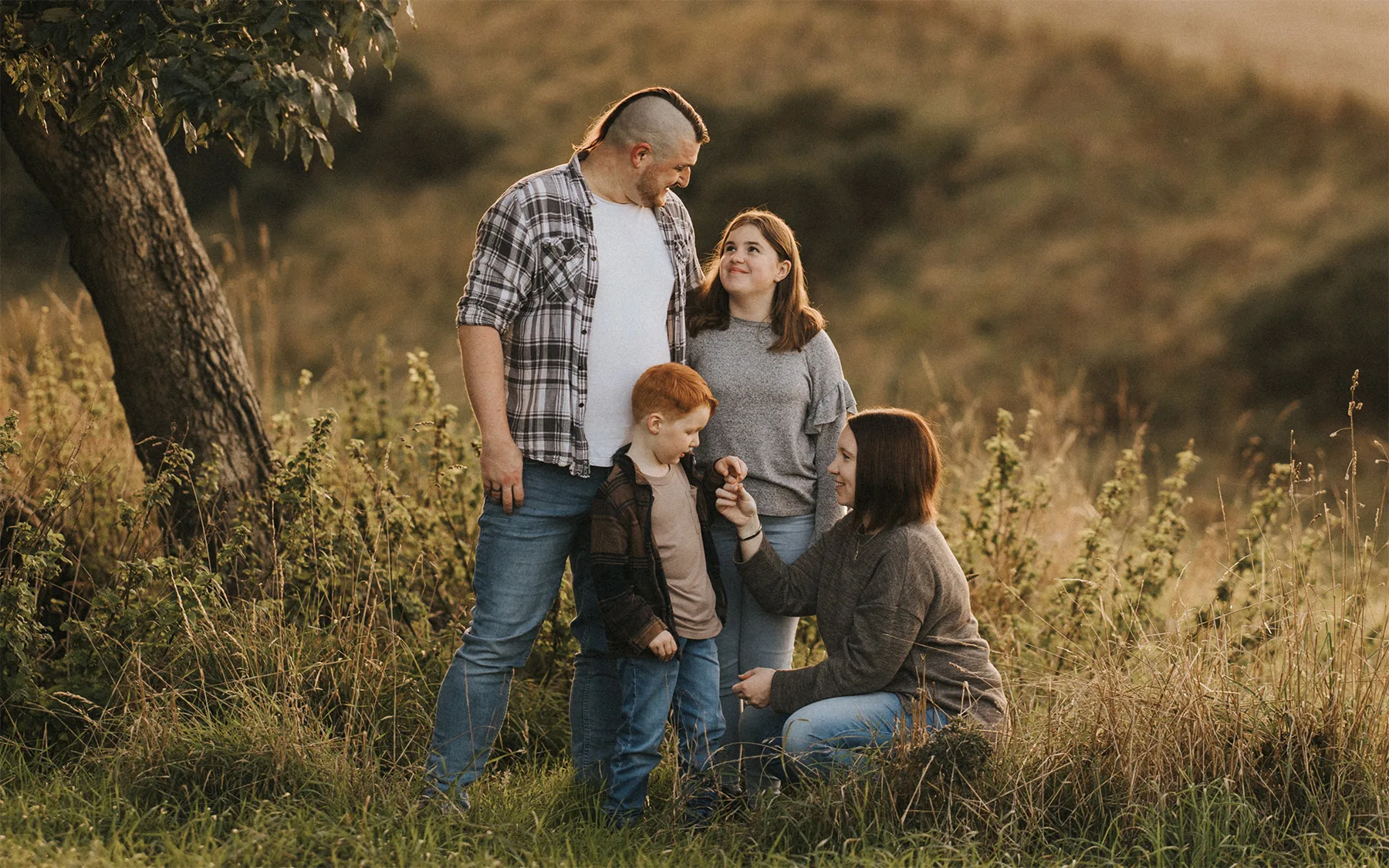 A family of four poses outdoors in tall grass at sunset, capturing a warm lifestyle moment. A man stands with a woman and boy, while another woman crouches, holding a flower to the boy, all smiling amid jeans, casual tops, a tree, and grassy hills. © Aimee Lince Photography - Wedding photographer in Lincolnshire, Yorkshire & Nottinghamshire