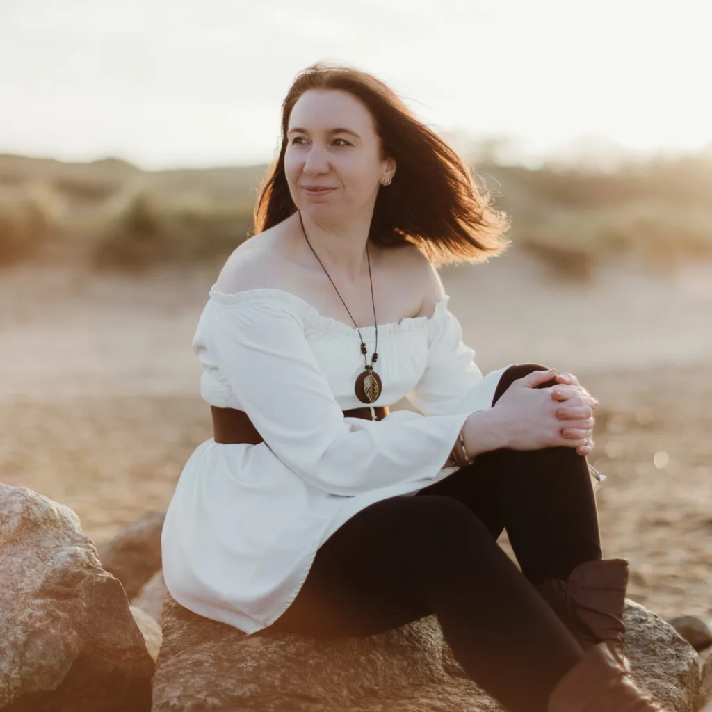 A woman with long brown hair wearing a white off-shoulder blouse and black trousers sits on rocks at a sandy beach, smiling and looking to the side as the sun sets behind her. © Aimee Lince Photography, wedding and lifestyle photographer in Lincolnshire