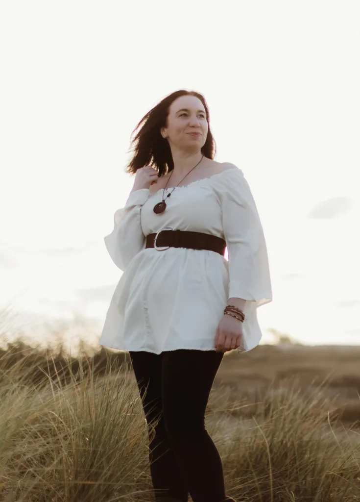 5.3. Photoshoot Web Res (Edited)-60-of-125-©-aimee-lince-photography-08-05-2026 A woman stands in tall grass under a bright sky, wearing a white tunic with a belt, black trousers, and bracelets. She looks into the distance with a calm expression, her hair blowing slightly in the wind. © Aimee Lince Photography, wedding and lifestyle photographer in Lincolnshire