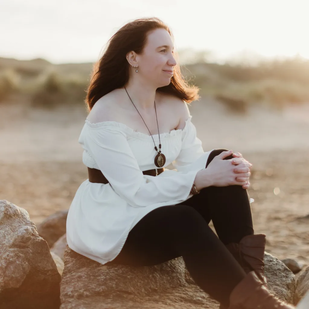 A woman in a white off-shoulder top and black trousers sits on rocks at a beach, looking into the distance. The sun sets behind her, casting a warm, golden glow over the sandy landscape. © Aimee Lince Photography, wedding and lifestyle photographer in Lincolnshire