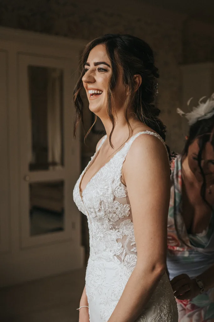 A smiling bride in a white lace wedding dress stands indoors, with another woman behind her helping adjust the dress. Warm light highlights the bride's joyful expression and elegant hairstyle. © Aimee Lince Photography, wedding and lifestyle photographer in Lincolnshire