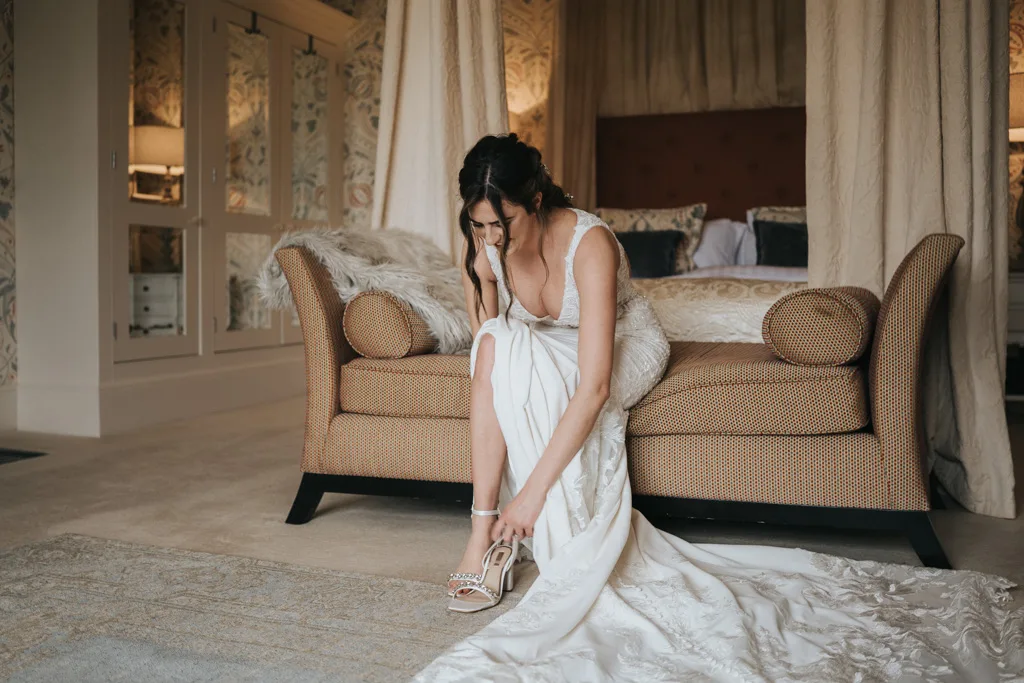 A bride in a white wedding dress sits on a tan sofa in a softly lit room, looking down as she fastens the strap on her high-heeled shoe. The train of her dress is spread out on the carpeted floor. © Aimee Lince Photography, wedding and lifestyle photographer in Lincolnshire