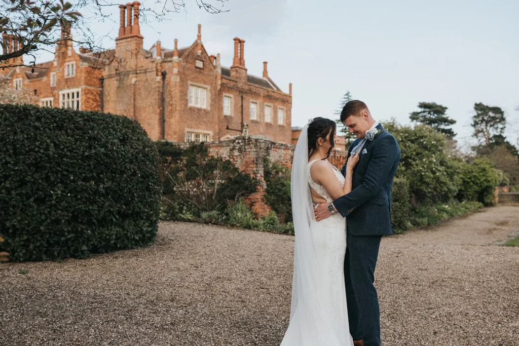 A bride and groom stand facing each other, embracing and smiling, outdoors on a gravel path with greenery and a large historic brick building in the background. © Aimee Lince Photography, wedding and lifestyle photographer in Lincolnshire