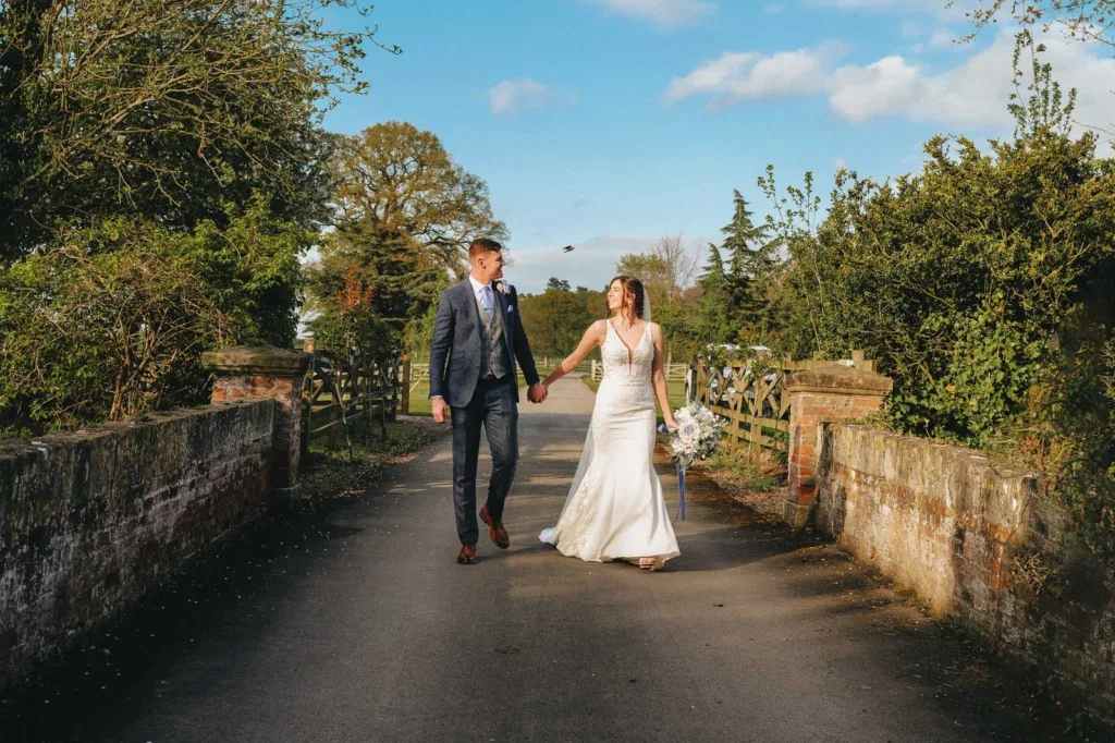 A bride in a white dress and a groom in a blue suit walk hand in hand down a country lane towards Hodsock Priory, surrounded by greenery and trees, under a bright blue sky with scattered clouds. © Aimee Lince Photography, wedding and lifestyle photographer in Lincolnshire