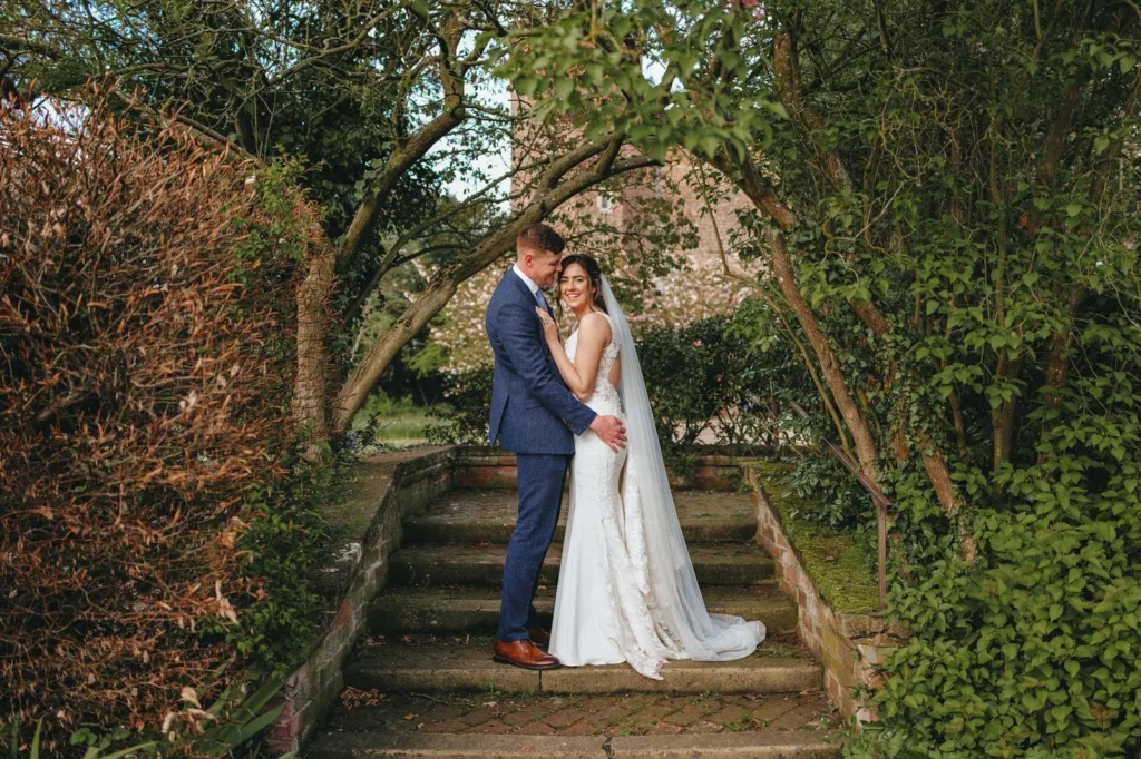 A bride in a white dress and veil stands with a groom in a blue suit on stone steps, surrounded by lush green trees and foliage, both smiling and embracing in a garden setting. © Aimee Lince Photography, wedding and lifestyle photographer in Lincolnshire