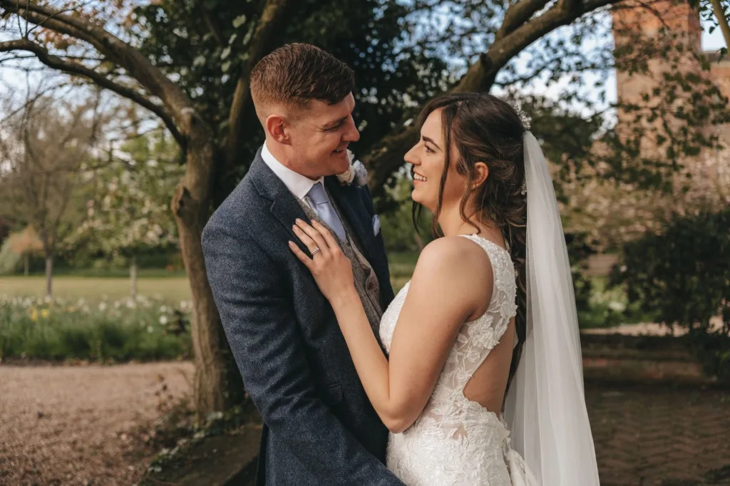 A bride and groom stand close, smiling lovingly at each other in an outdoor garden setting. The bride wears a white lace dress and veil, while the groom is in a blue suit. Trees and greenery are visible in the background. © Aimee Lince Photography, wedding and lifestyle photographer in Lincolnshire