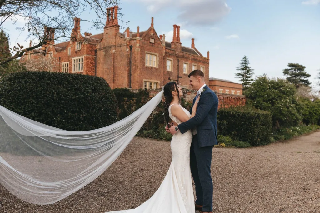 A bride and groom stand facing each other, smiling, in front of a large red-brick mansion. The bride’s veil flows in the wind, and the outdoor scene is framed by greenery and a partly cloudy sky. © Aimee Lince Photography, wedding and lifestyle photographer in Lincolnshire