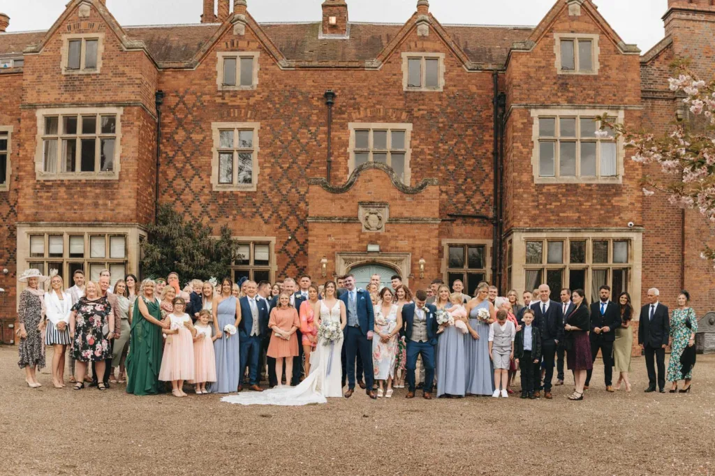 A large wedding party poses in front of a historic brick mansion, with the bride and groom in the centre surrounded by family and friends dressed in formal attire. © Aimee Lince Photography, wedding and lifestyle photographer in Lincolnshire