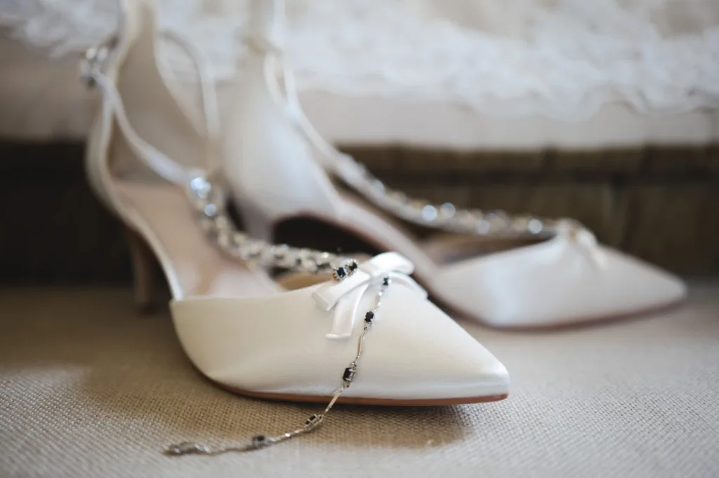 A pair of elegant white satin high-heeled shoes with pointed toes and small bows, decorated with a delicate beaded necklace, resting on a beige fabric surface. © Aimee Lince Photography, wedding and lifestyle photographer in Lincolnshire