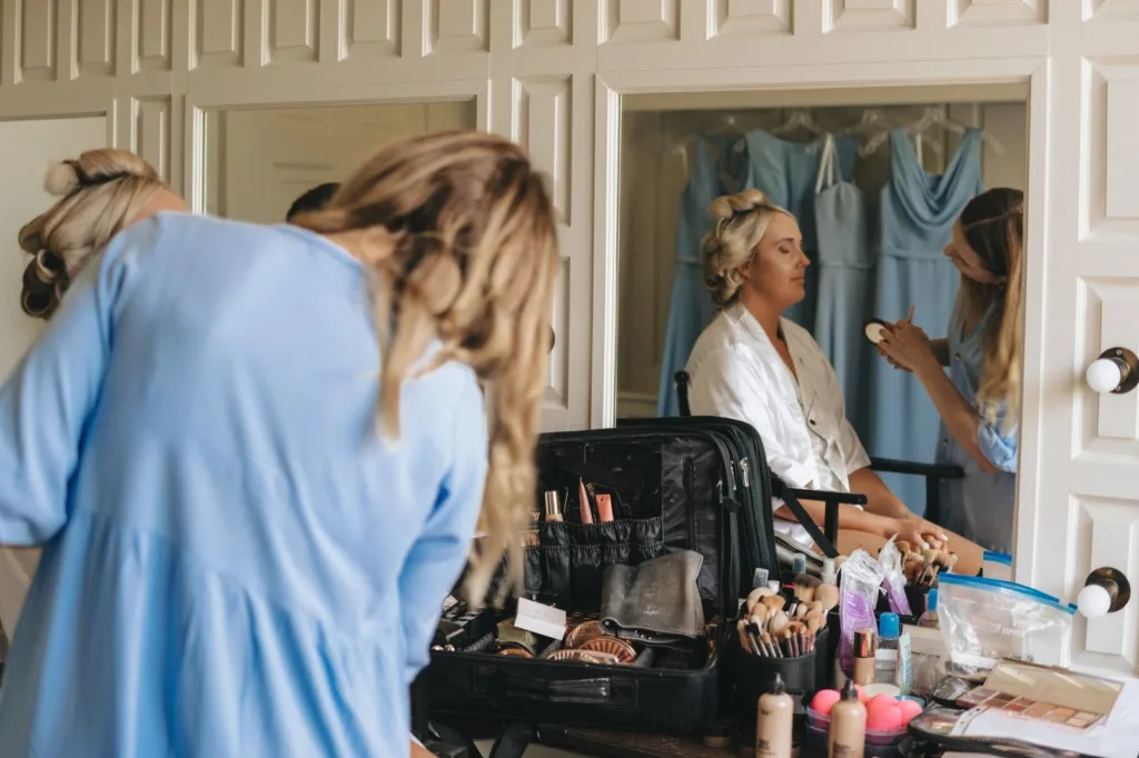 Two women in a changing room; one is applying make-up to the other, who is seated in front of a mirror. The table is covered with make-up items, and light blue dresses hang in the background. © Aimee Lince Photography, wedding and lifestyle photographer in Lincolnshire