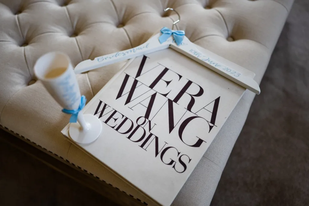 A book titled "Vera Wang on Weddings" rests on a tufted beige cushion, alongside a white clothes hanger labelled "Bridesmaid" and "10th June 2023" in blue, and a rolled paper tied with a blue ribbon. © Aimee Lince Photography, wedding and lifestyle photographer in Lincolnshire