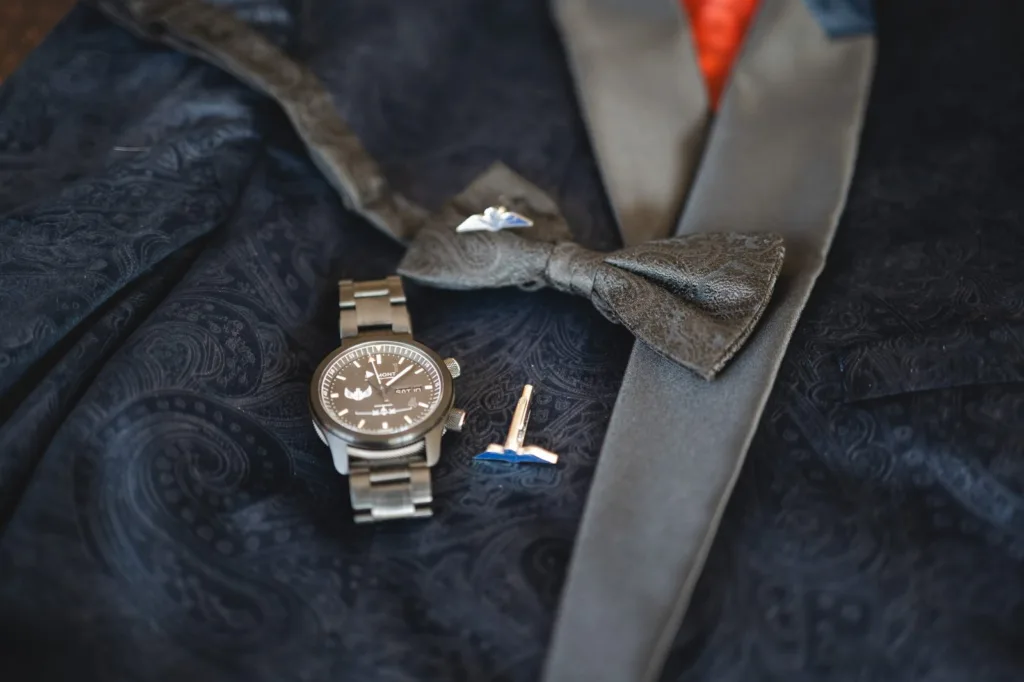 A close-up of a navy paisley suit jacket with a grey patterned bow tie, a silver wristwatch, and two cufflinks placed on top. The jacket has a dark satin lapel and an orange inner lining peeking through. © Aimee Lince Photography, wedding and lifestyle photographer in Lincolnshire