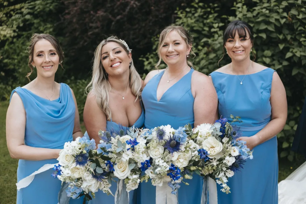 Four women in matching blue dresses stand outdoors, smiling and holding large bouquets of white and blue flowers, with greenery in the background. © Aimee Lince Photography, wedding and lifestyle photographer in Lincolnshire