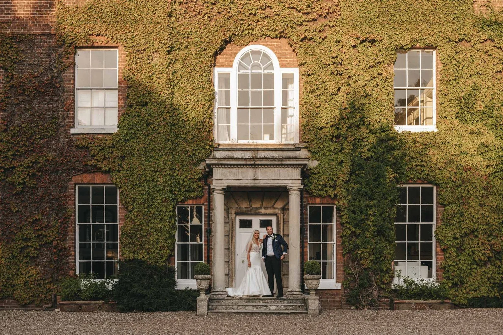 A bride and groom stand together on the front steps of a large, ivy-covered brick building with tall windows and a grand entrance. © Aimee Lince Photography, wedding and lifestyle photographer in Lincolnshire