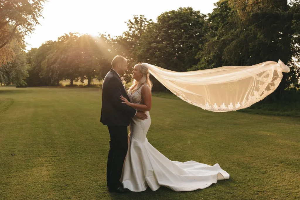 A bride and groom stand on a grassy field at sunset. The bride wears a white gown with a long veil flowing in the breeze, whilst the groom wears a dark suit. They look at each other lovingly, surrounded by trees. © Aimee Lince Photography, wedding and lifestyle photographer in Lincolnshire