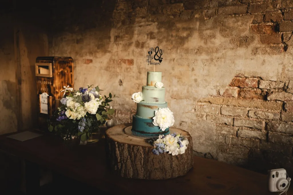 A three-tiered blue and teal wedding cake with white flowers sits on a wooden slice, next to a bouquet and a vintage postbox, against a rustic brick wall. © Aimee Lince Photography, wedding and lifestyle photographer in Lincolnshire