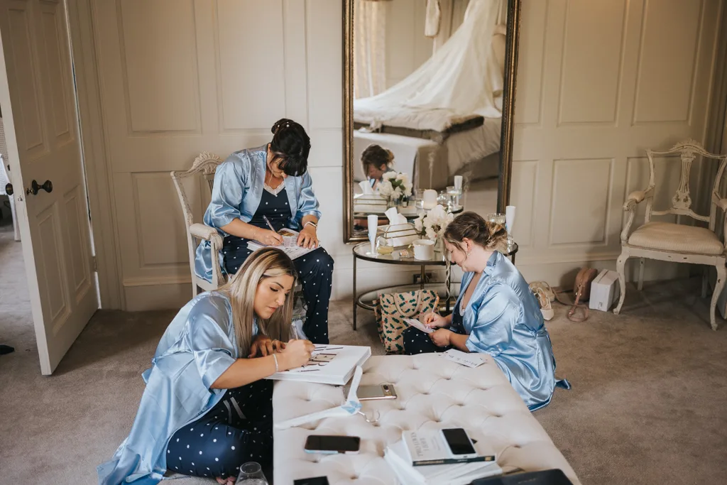 Three women wearing light blue satin dressing gowns sit in a cosy, elegant room, writing or drawing in notebooks. A large mirror, chairs, and a table with flowers and drinks are visible in the softly lit space. © Aimee Lince Photography, wedding and lifestyle photographer in Lincolnshire