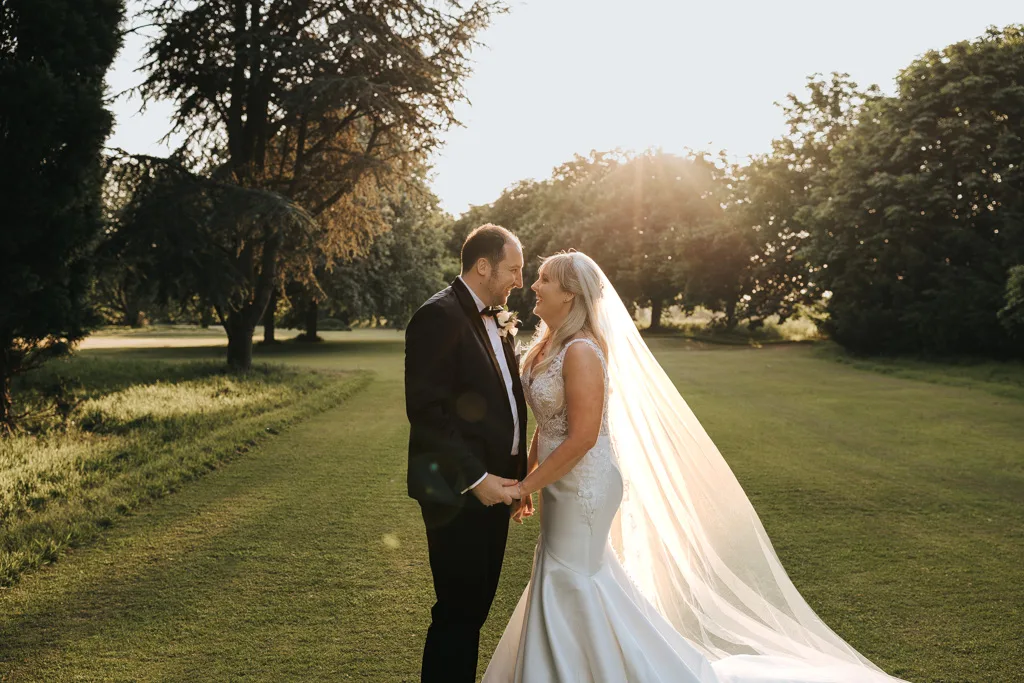 A bride and groom stand facing each other on a grassy lawn, holding hands and smiling, with sunlight streaming through trees behind them. The bride wears a long white dress and veil, and the groom is in a black suit. © Aimee Lince Photography, wedding and lifestyle photographer in Lincolnshire