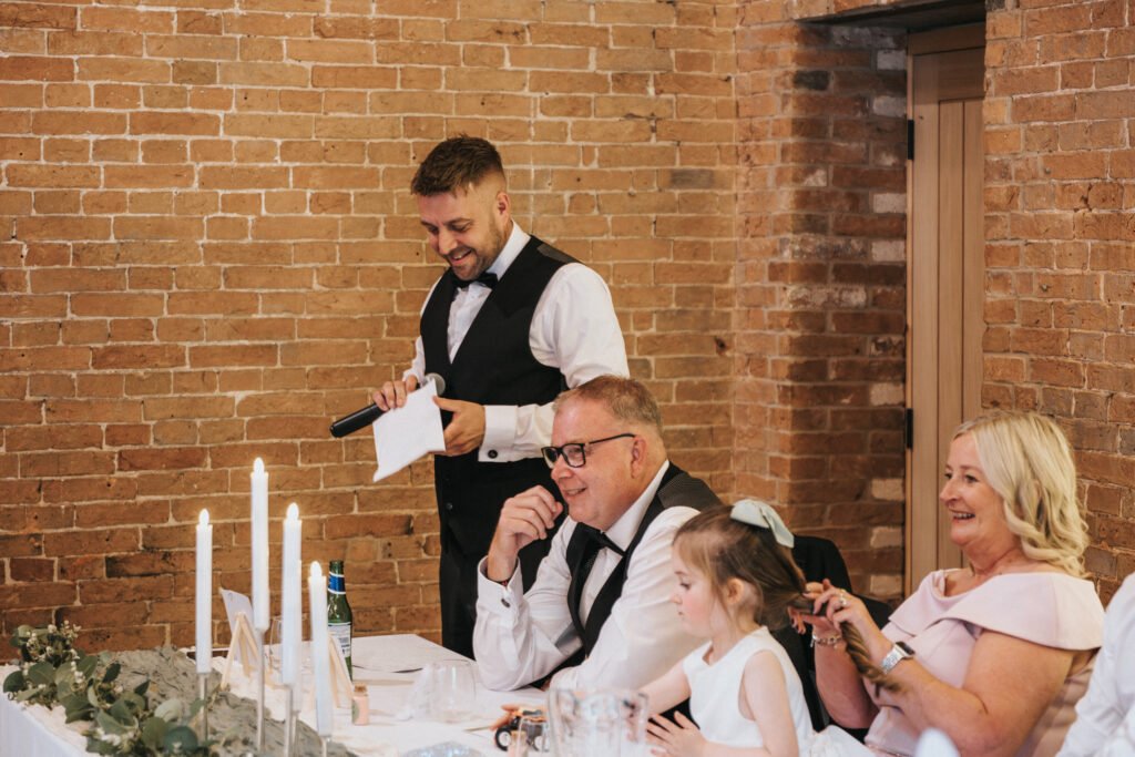 A man in a waistcoat and bow tie stands smiling with a microphone and paper, giving a speech at an Elsham Hall table. Beside him, guests beam as a wedding photographer captures the moment amid white candles and greenery. © Aimee Lince Photography, Lincolnshire photographer