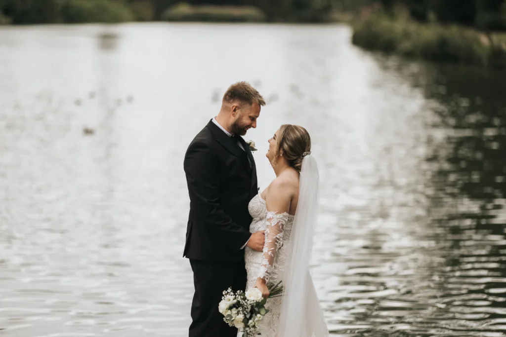 Award winning wedding image. The Guild of Wedding Photographers, bronze award. A bride in a white lace wedding dress and a groom in a dark suit walk hand-in-hand down a garden path, as their wedding photographer captures the moment amid manicured trees, lush grass, and a brick wall under a cloudy sky. © Aimee Lince Photography, Lincolnshire photographer