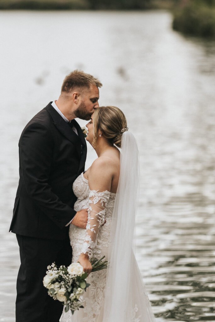 A groom in a black suit gently kisses the forehead of his bride, her lace gown and long veil trailing behind, as they stand by a calm lake at Elsham Hall. Captured by a talented wedding photographer, greenery lines the water’s edge. © Aimee Lince Photography, Lincolnshire photographer