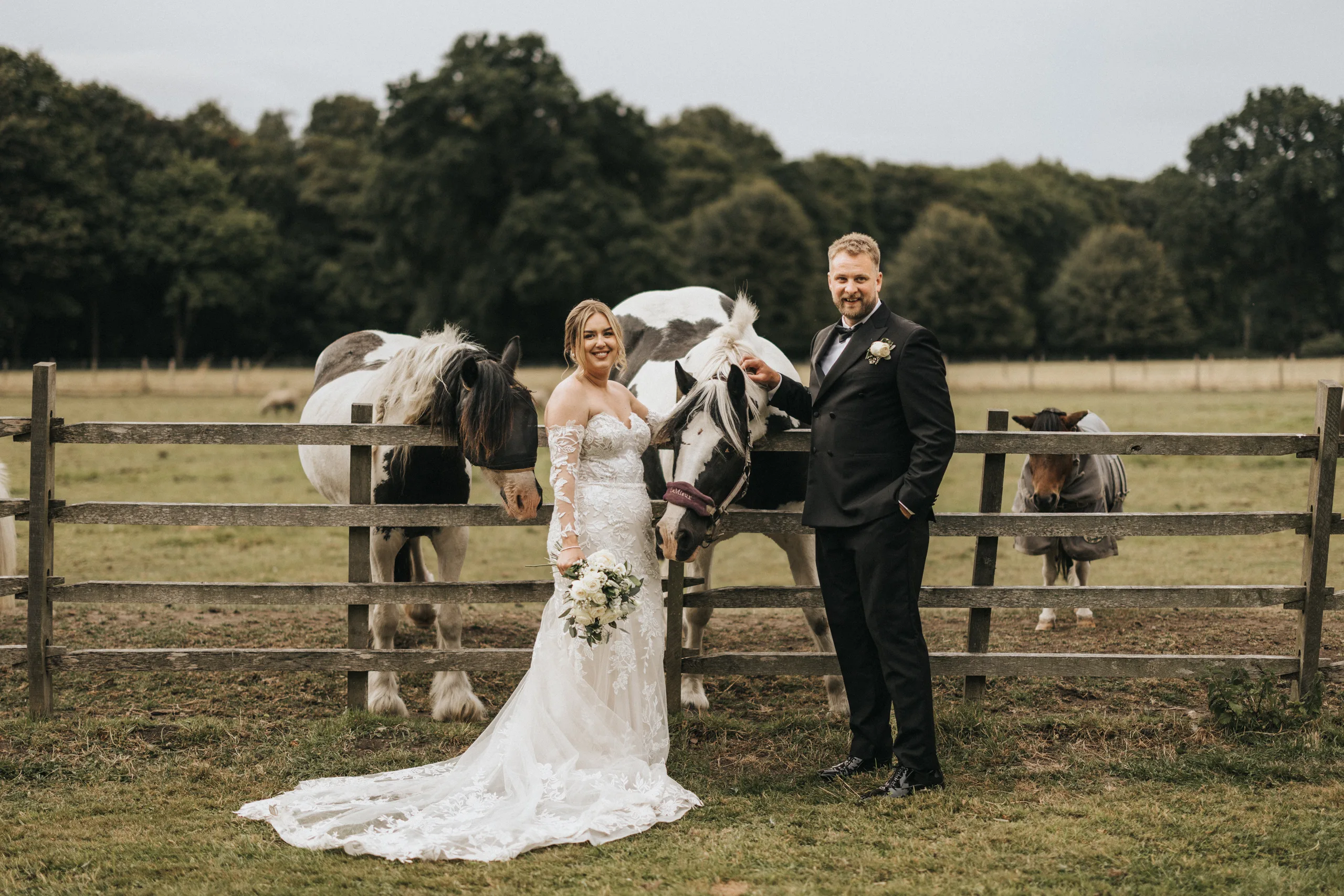 A bride in a white gown and a groom in a black suit stand smiling by a wooden fence at Elsham Hall with two horses, one of which the groom is stroking. Trees and grassy fields fill the background, captured beautifully by their wedding photographer. © Aimee Lince Photography, Lincolnshire photographer