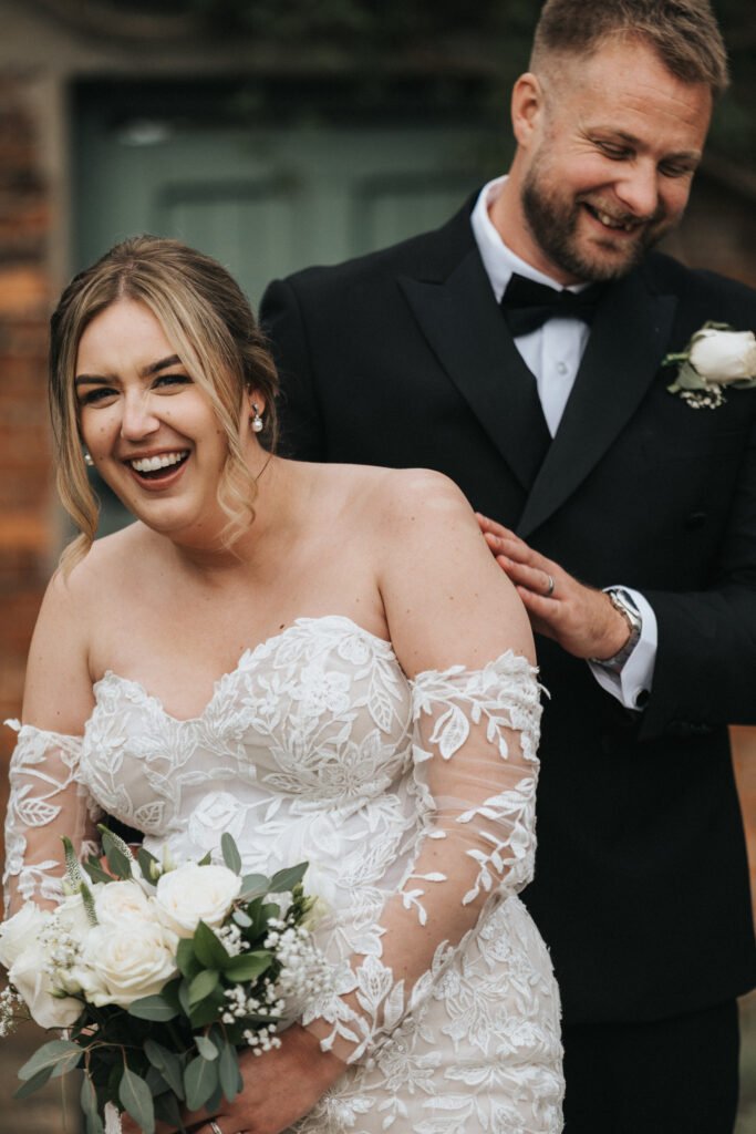 A smiling bride in an off-shoulder lace wedding dress holds a white rose bouquet. Her groom, in a black tuxedo with a white buttonhole, stands behind her at Elsham Hall, both joyful and relaxed as their wedding photographer captures the moment outdoors. © Aimee Lince Photography, Lincolnshire photographer