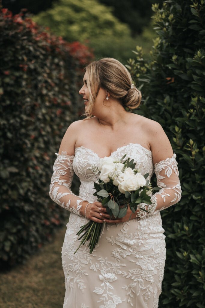 A bride in an off-the-shoulder, lace wedding dress holds a white rose bouquet and smiles to her left. Her hair is styled in a low bun as she stands outdoors between tall green hedges at Elsham Hall, captured by a skilled wedding photographer. © Aimee Lince Photography, Lincolnshire photographer