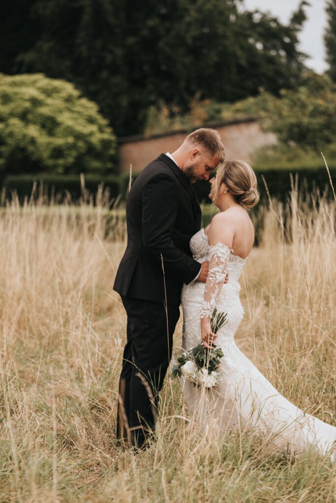 A bride in a lace wedding dress and a groom in a black suit stand close together in a tall grassy field at Elsham Hall, touching foreheads and smiling softly. Captured by a wedding photographer, the bride holds a bouquet of white flowers. © Aimee Lince Photography, Lincolnshire photographer