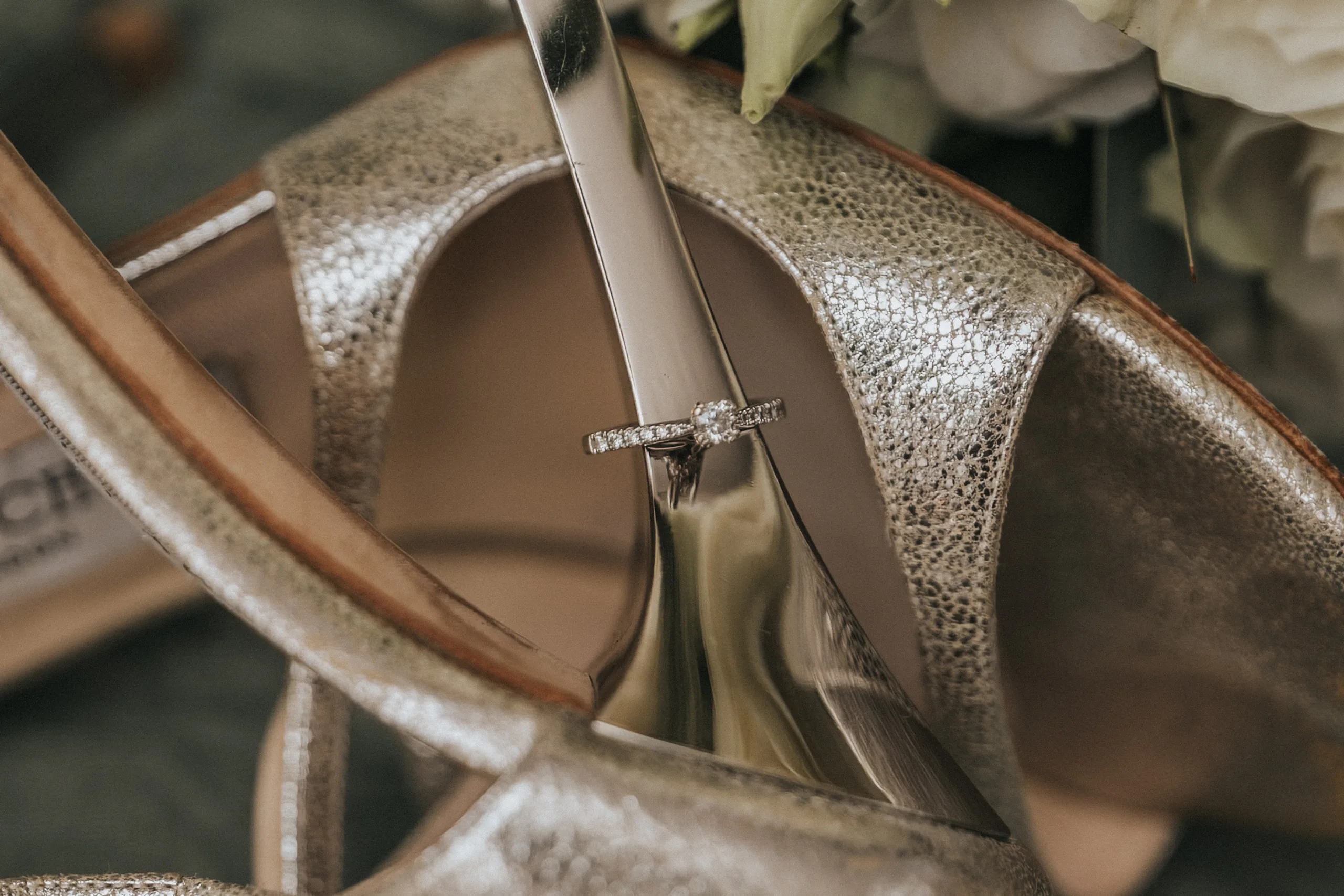 A close-up of a silver diamond engagement ring balanced on the heel of metallic gold high-heeled shoes, their textured, shimmering surface glowing. Soft white flowers blur in the background—perfect for a wedding photographer at Elsham Hall. © Aimee Lince Photography, Lincolnshire photographer