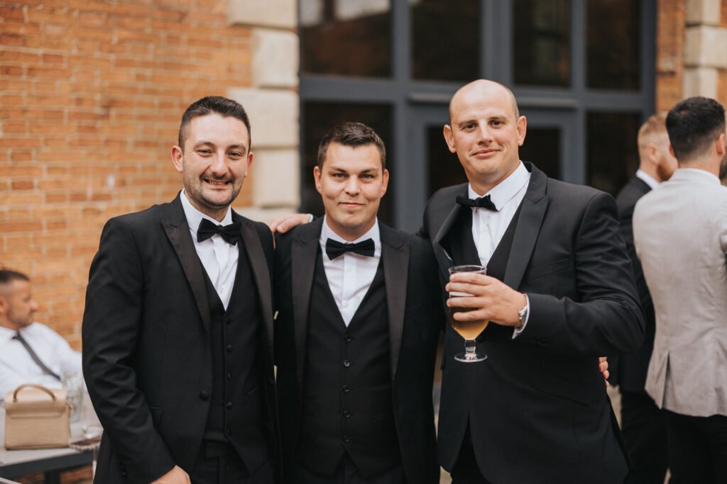 Three men in black dinner jackets with bow ties stand close together, smiling at the camera. One man holds a drink. Captured by a wedding photographer at Elsham Hall, they pose outdoors before a brick wall and large windows, guests blurred behind them. © Aimee Lince Photography, Lincolnshire photographer