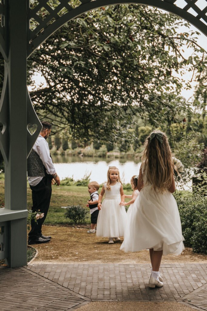 Three children in white dresses and a toddler in a striped outfit stand outdoors near a lake at Elsham Hall. An adult in a waistcoat, shirt, and tie stands to the left beneath an arched wooden structure—perfect for any wedding photographer’s portfolio. © Aimee Lince Photography, Lincolnshire photographer