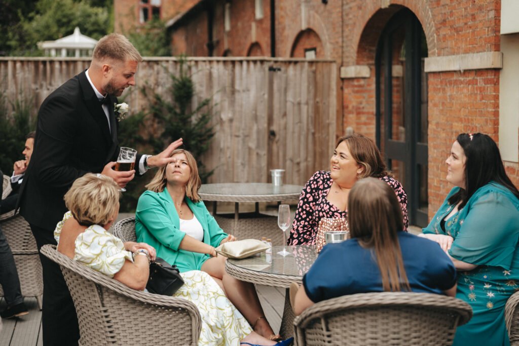 A man in a black suit stands by a patio table, smiling and gesturing as he talks to five seated women at Elsham Hall. The women, dressed in colourful dresses, sit on wicker chairs outdoors against a brick building as drinks rest on the table—a perfect scene for a wedding photographer. © Aimee Lince Photography, Lincolnshire photographer