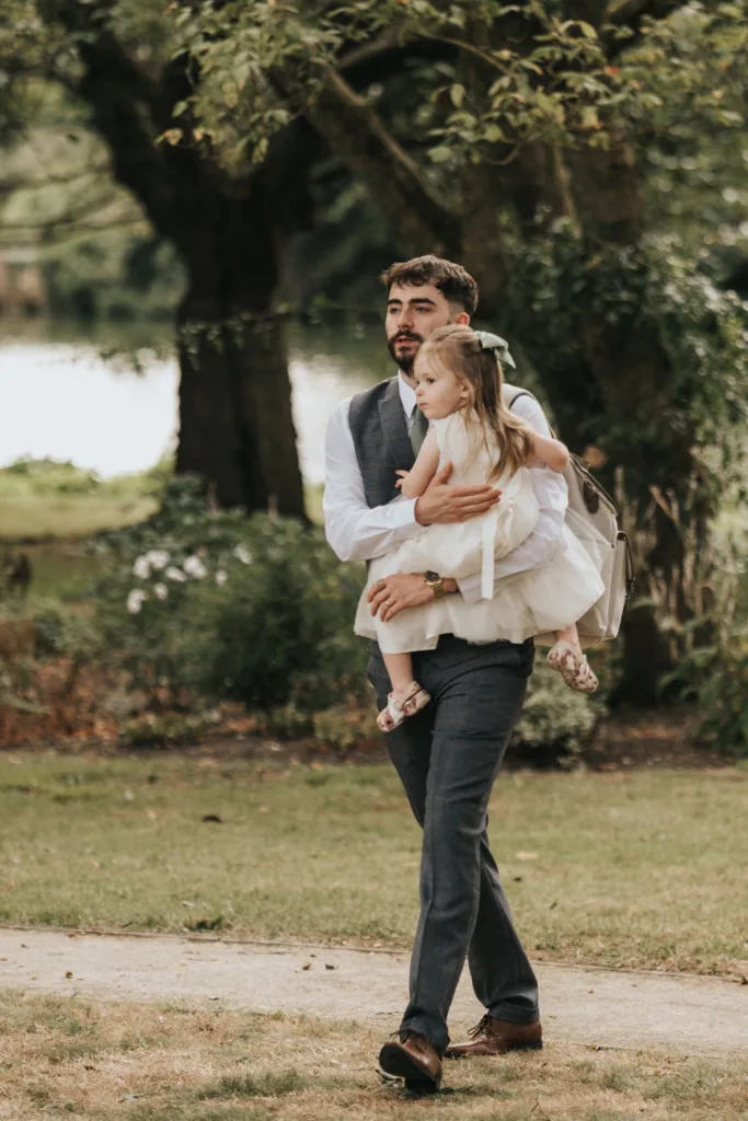 A bearded man in a blue suit and waistcoat carries a young girl in a white dress and sandals. Walking outdoors at Elsham Hall, they’re surrounded by green grass, trees, and soft sunlight—an ideal moment for a wedding photographer to capture. © Aimee Lince Photography, Lincolnshire photographer