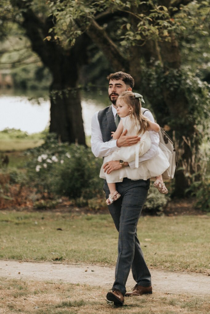 A bearded man in a blue suit and waistcoat carries a young girl in a white dress and sandals. Walking outdoors at Elsham Hall, they’re surrounded by green grass, trees, and soft sunlight—an ideal moment for a wedding photographer to capture. © Aimee Lince Photography, Lincolnshire photographer